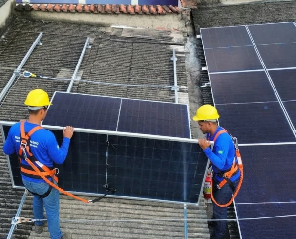 Instaladores da ASG Energia, com capacete e cinto de segurança, carregando placa solar